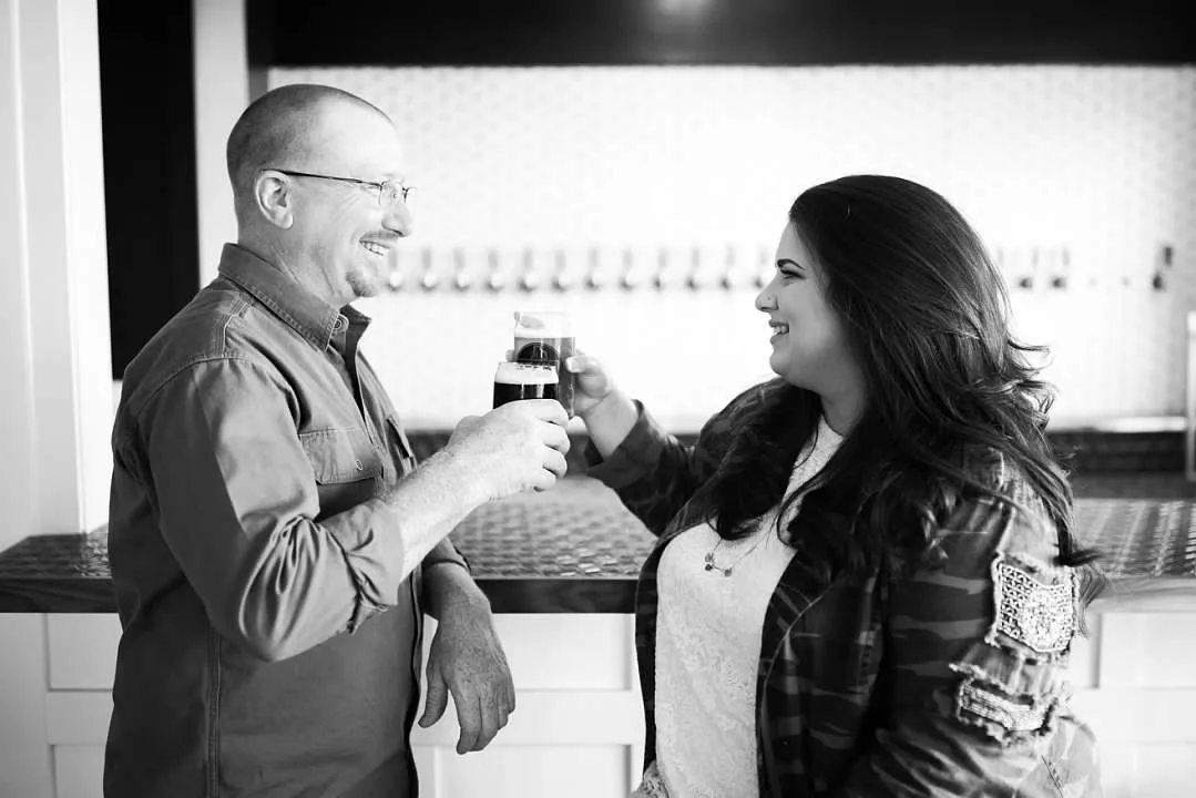 Black and white photo of two people at the bar, clinking a glass together, with the tap wall lined up behind them.