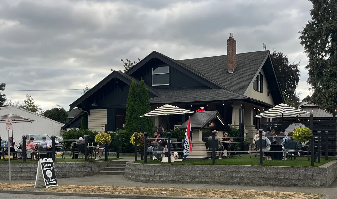 Fill's Growlers on an overcast evening: a black and cream 1914 craftsman house with guests at tables under striped umbrellas on the front lawn.